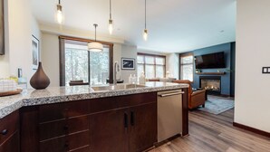 A modern kitchen with granite countertops, dark wood cabinets, and a built-in dishwasher. There are pendant lights, a large window with blinds, and a fireplace in the background. - Luxurious Home on Sierra Star Golf Course. Balcony & Gas Grill, Washer/Dryer (Mammoth Lakes)