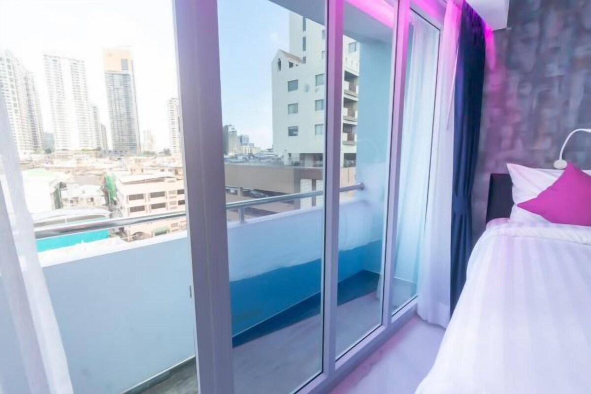 A hotel room with a balcony, large windows, and a bed with white bedding and a pink pillow.