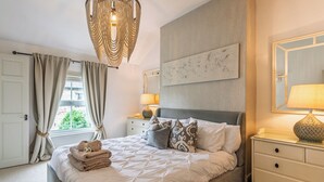 Uma cama bem arrumada com colcha de padrão geométrico, um abajur, um espelho fixado na parede e vista para o exterior através de uma janela com cortinas.
