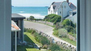 A coastal view from a window overlooking a road leading to a white house with a curved roof, a stone wall, and a grassy area with shrubs.