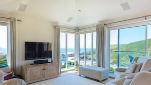 A spacious living room with a large flat-screen TV, a comfortable sofa, and a view of the ocean through large windows.