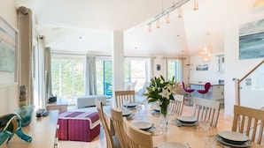 A spacious dining area with a wooden table set for six, surrounded by wooden chairs. There is a vase with white flowers on the table, and the room has large windows allowing natural light.