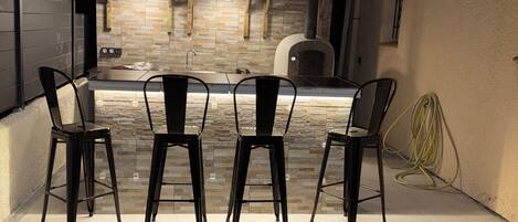 An outdoor kitchen with a stone backsplash, hanging pendant lights, and four metal chairs.