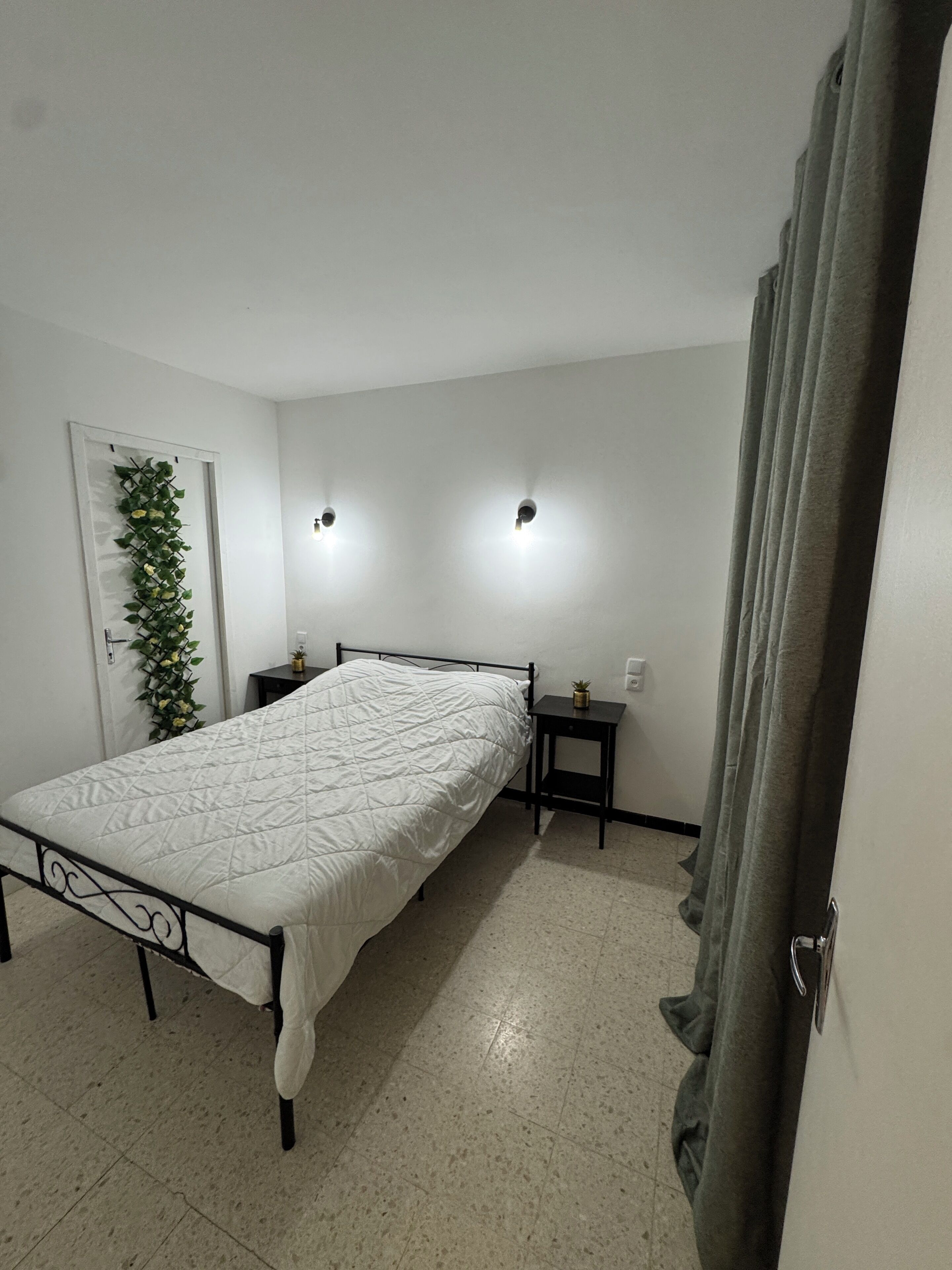 A bedroom with a bed, a nightstand, a wall-mounted light, and a door with a curtain.