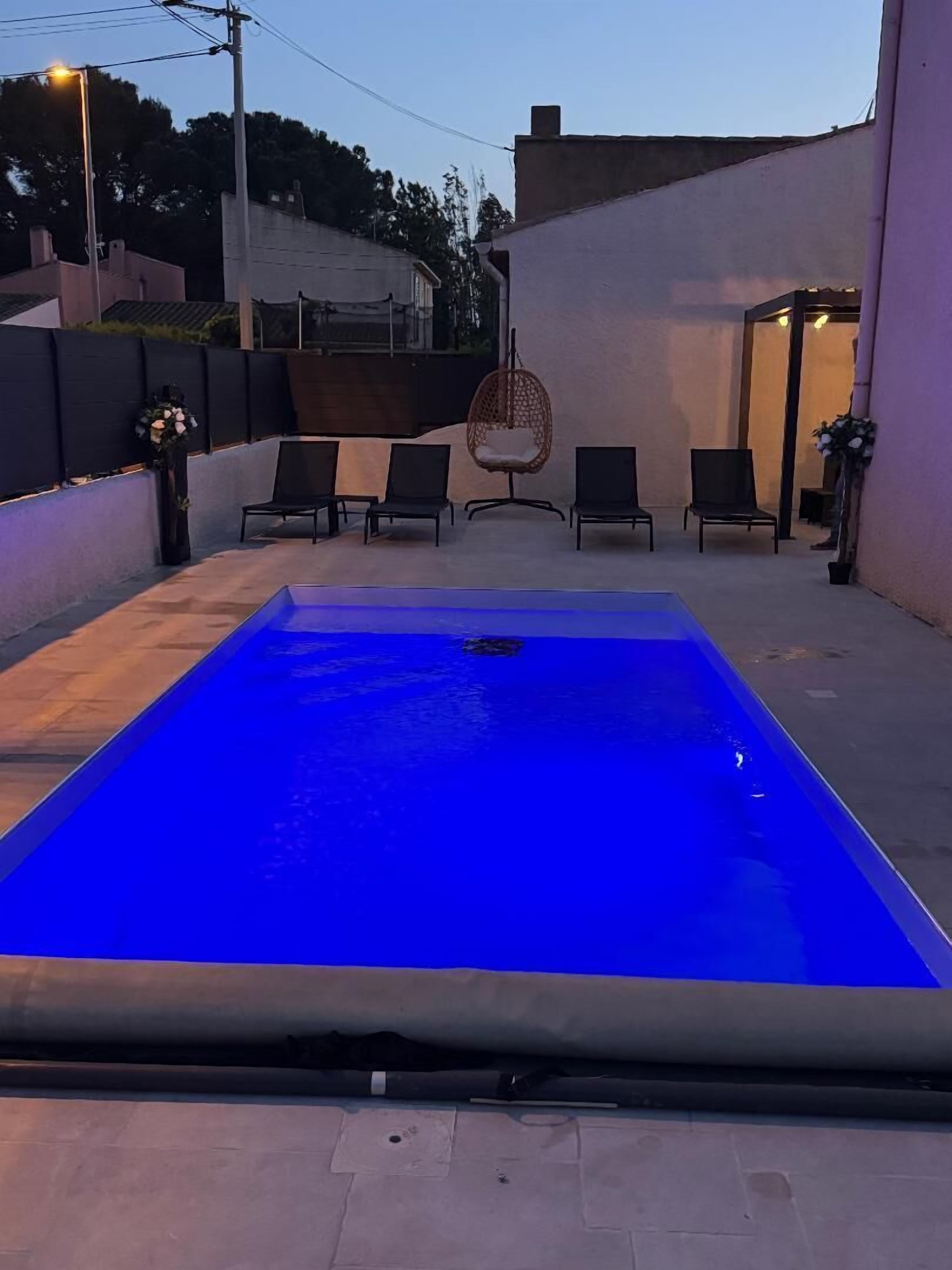 Una piscina rectangular con un ligero tono azul, rodeada por un patio empedrado con sillas reclinables negras y una mecedora colgante.