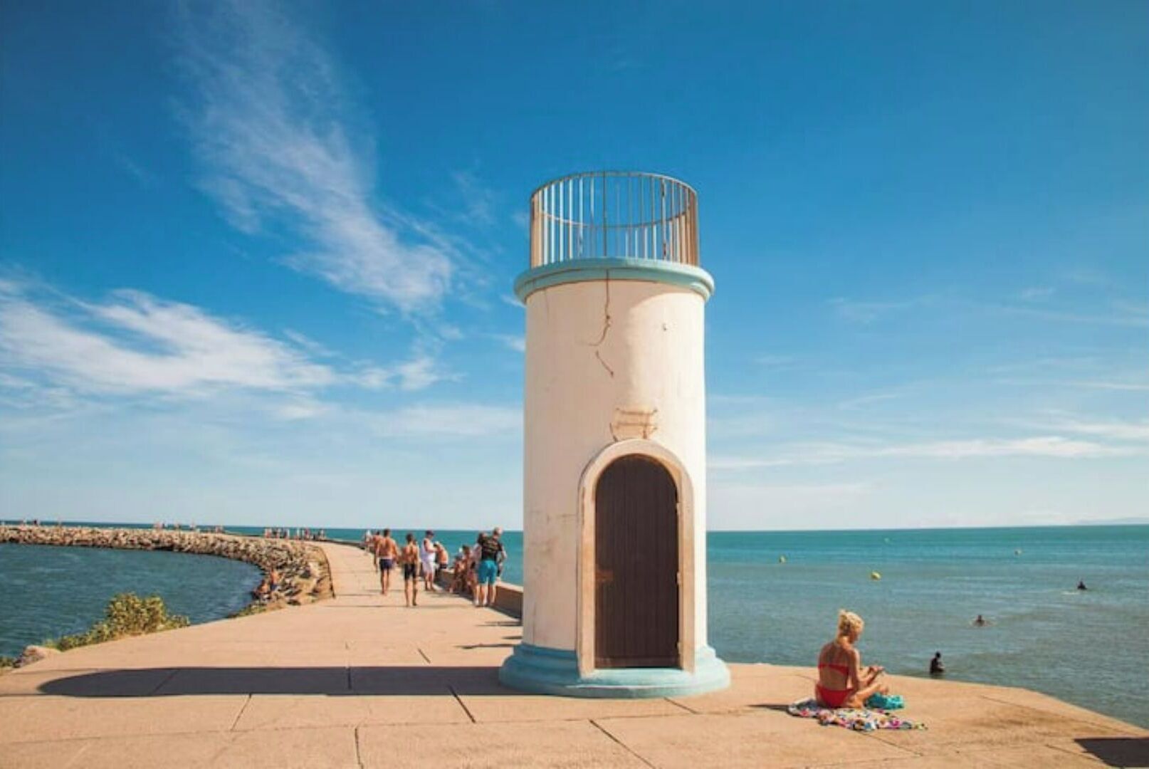 A coastal walkway with a cylindrical lighthouse structure, a stone pier, and a beach with people and water.