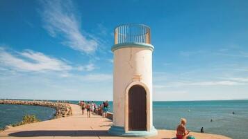 A coastal walkway with a cylindrical lighthouse structure, a stone pier, and a beach with people and water.