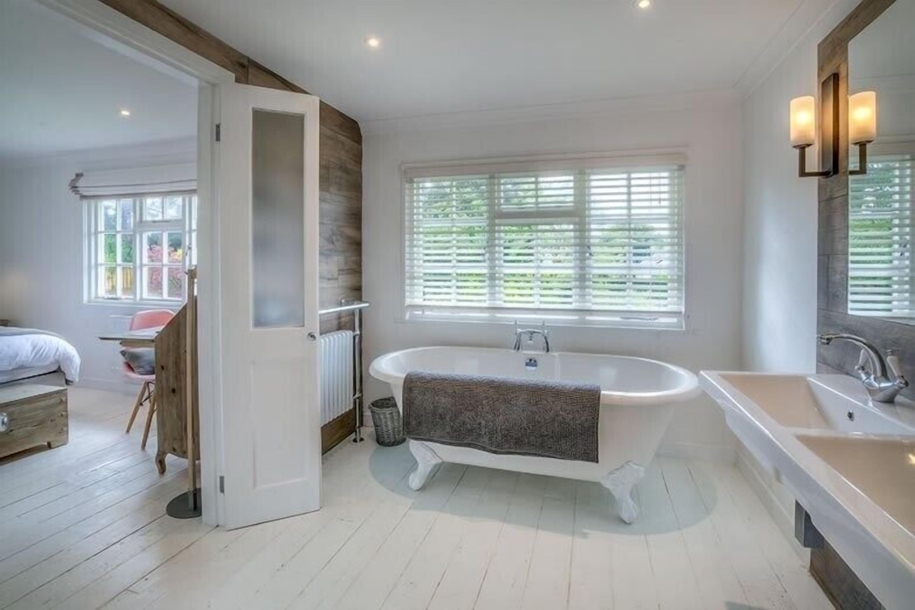 Une salle de bain moderne avec une baignoire sur pieds, une grande fenêtre avec stores et un miroir agrémenté de luminaires fixés au mur.