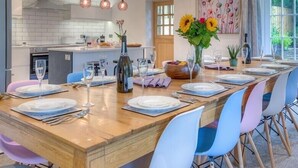 A dining area with a long wooden table set for six, surrounded by colorful chairs, and a kitchen in the background. - The Coach House (Pwllheli)
