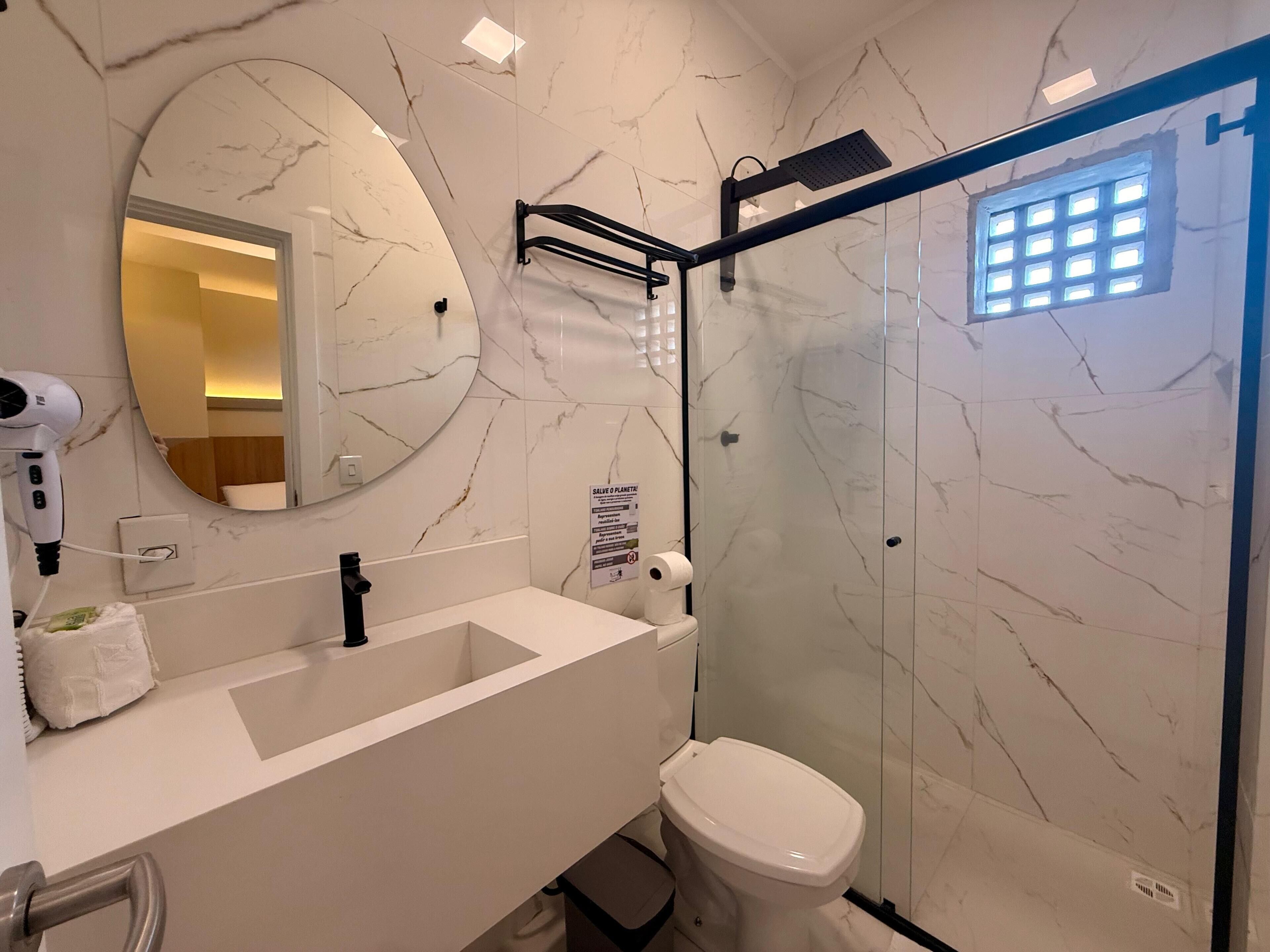 A modern bathroom with a marble backsplash, a round mirror, a white sink, a black faucet, a white toilet, and a glass-enclosed shower area.