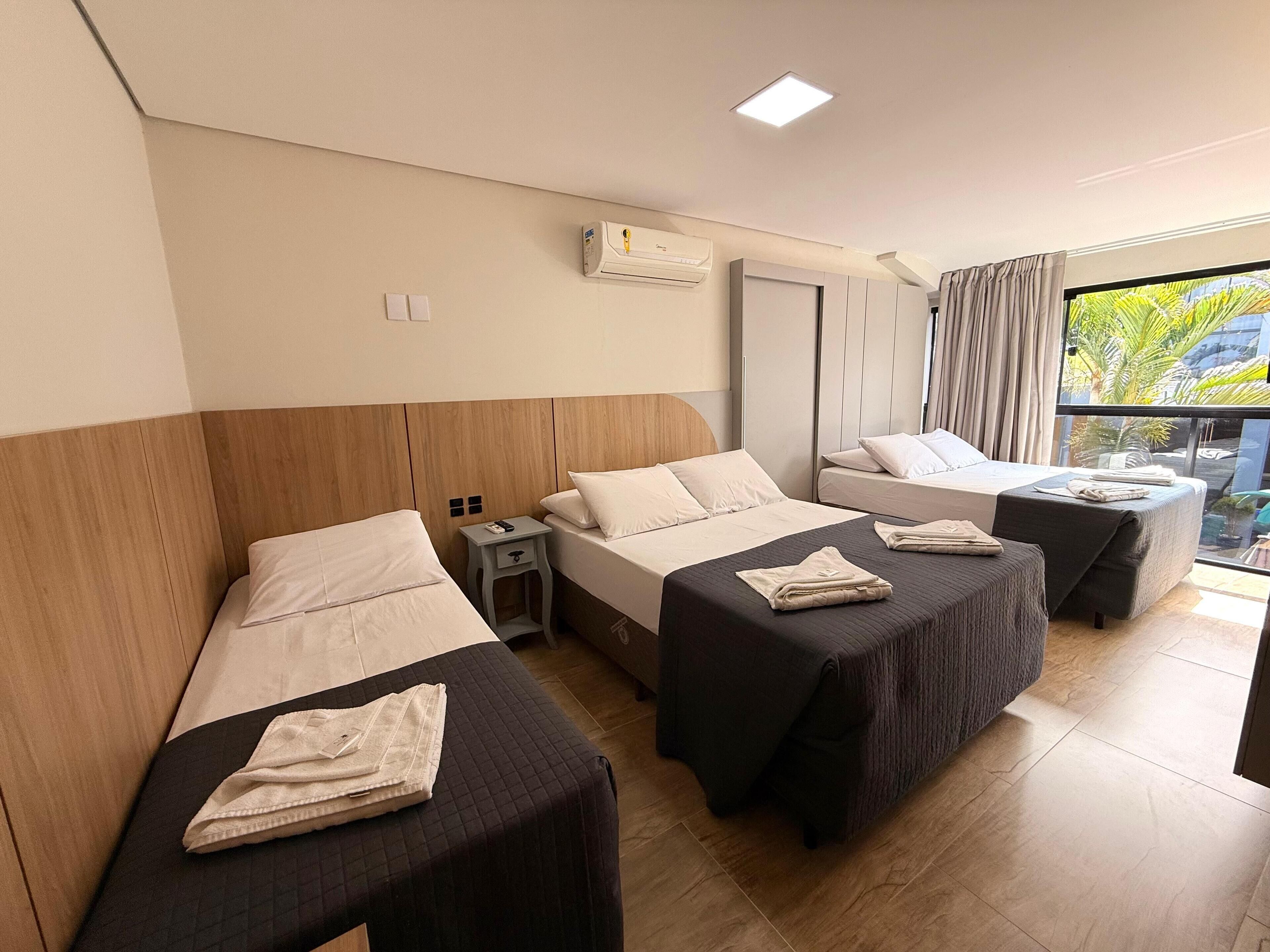 A hotel room with three beds, a small table, and a window with a view of palm trees.