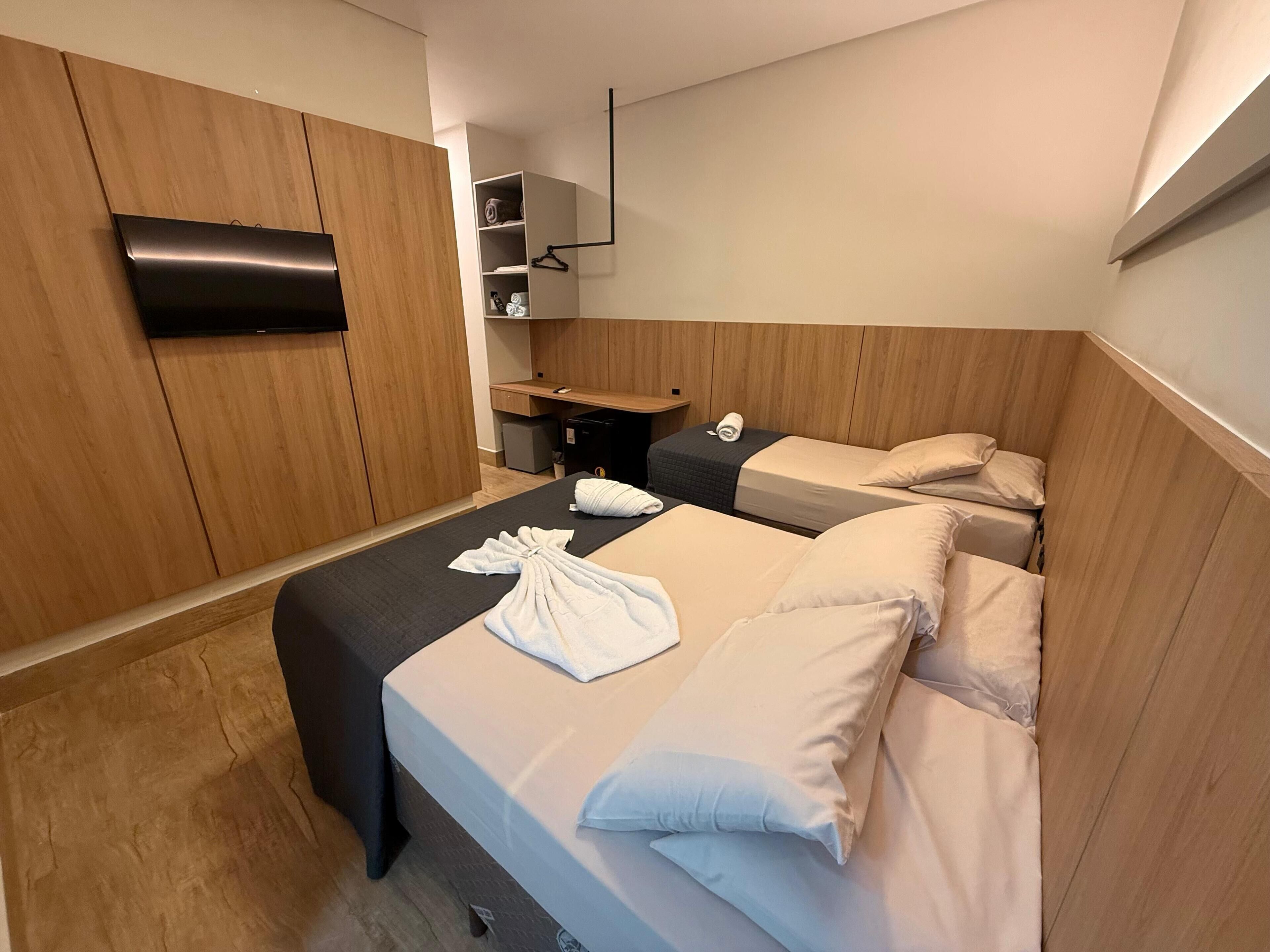 A hotel room with two beds, a wooden wardrobe, a desk with a chair, and a small shelf with towels.