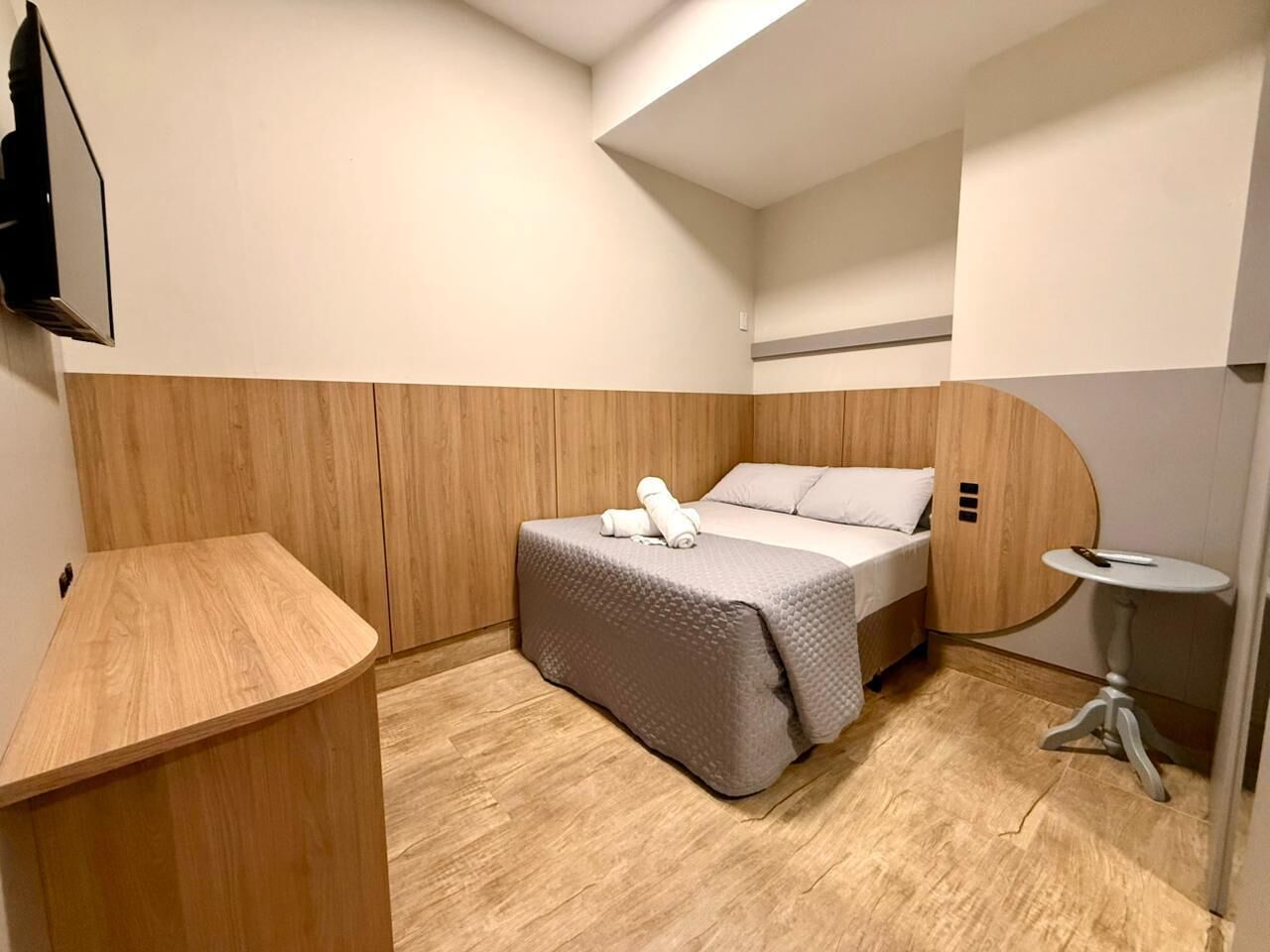 Une chambre d’hôtel compacte comprenant un lit, une petite table de chevet, un téléviseur fixé au mur et un décor avec des panneaux en bois.