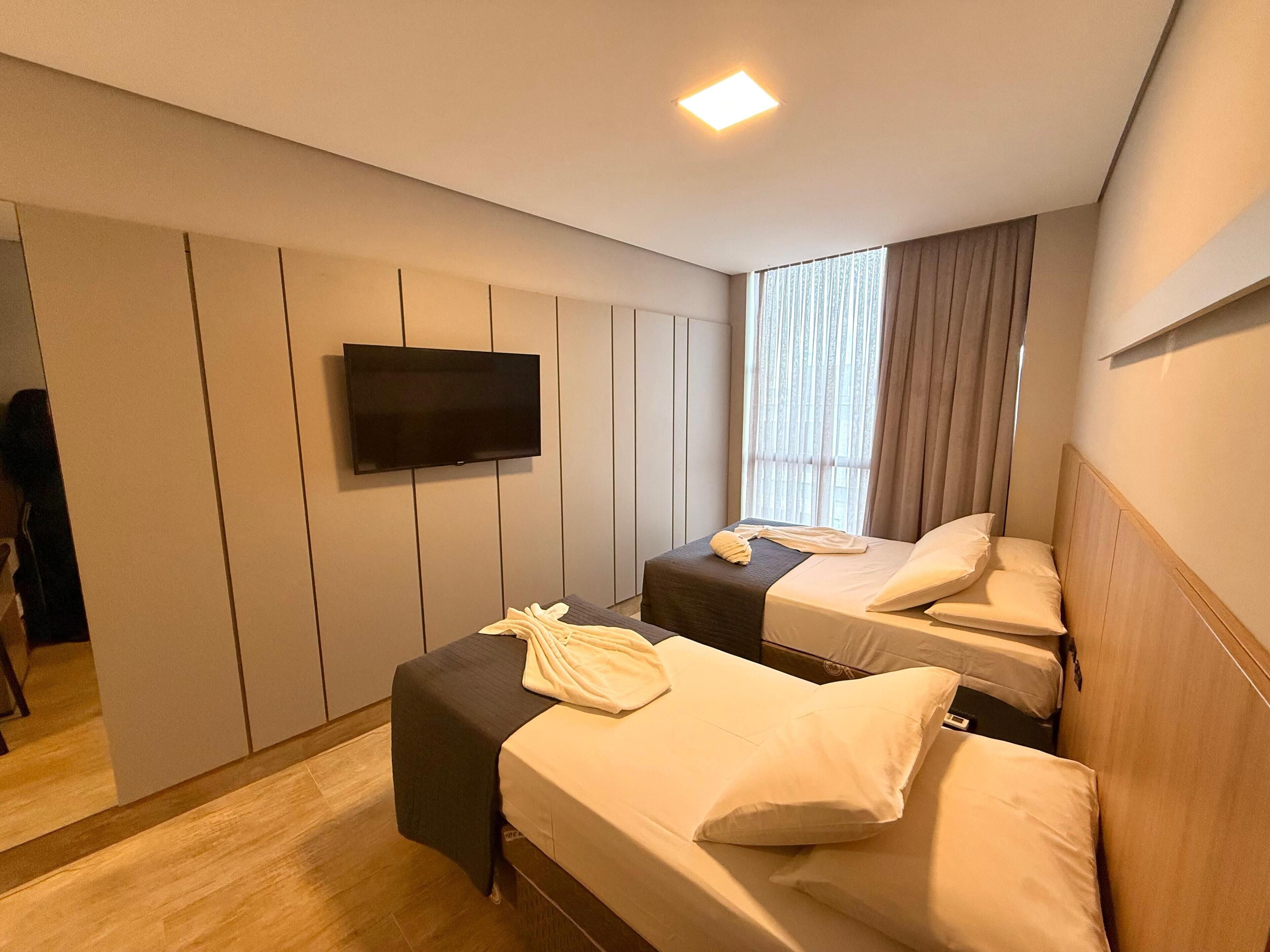 A hotel room with two single beds, a flat-screen TV mounted on the wall, a window with curtains, and a wooden floor.
