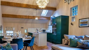 A spacious living area with a dining table, blue upholstered chairs, a sofa, and a coffee table. A kitchen with green cabinets and modern appliances is visible in the background. - A luxury barn conversion close to Grizedale Forest & Hawkshead village (Satterthwaite (Nr. Hawkshead))