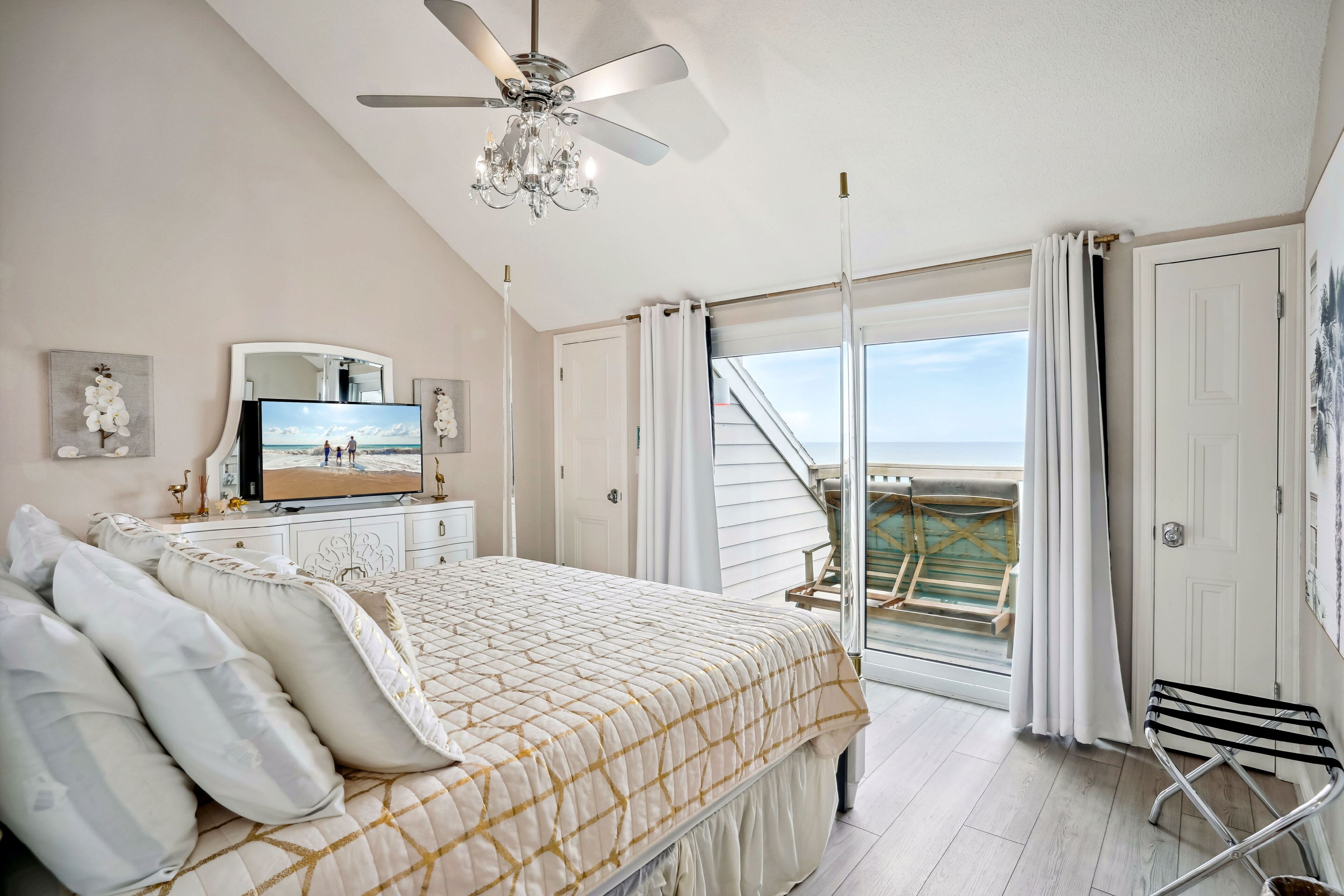 A bedroom with a bed, a ceiling fan, a TV, and a balcony with a view of the ocean.