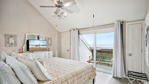 Un dormitorio con cama, ventilador de techo, televisor y balcón con vista al océano.