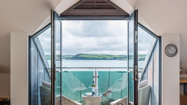 A room with a glass door leading to a balcony with a view of the sea and hills. - Upper Deck (Wadebridge)