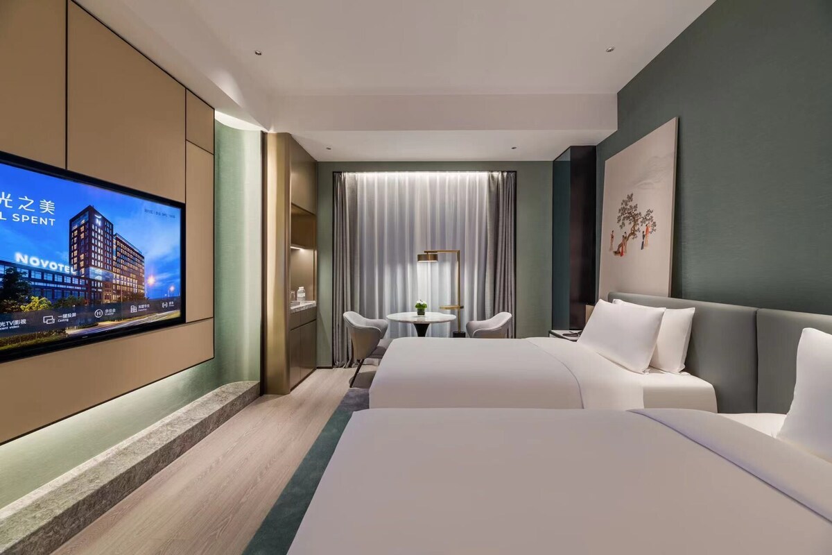 Quarto de hotel com uma cama grande, uma escrivaninha com cadeira, televisão na parede e janela com cortinas.