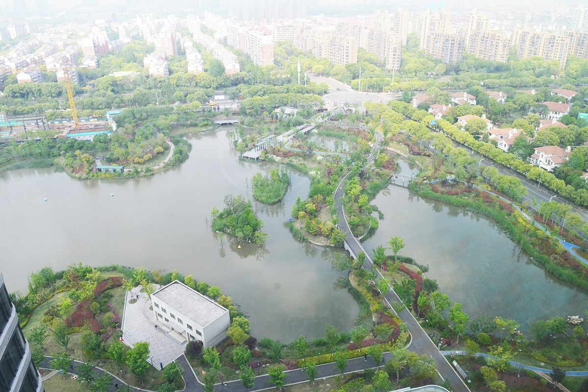 Vista aérea de um parque à beira-rio, com caminhos para pedestres, vegetação e edifícios residenciais ao fundo.