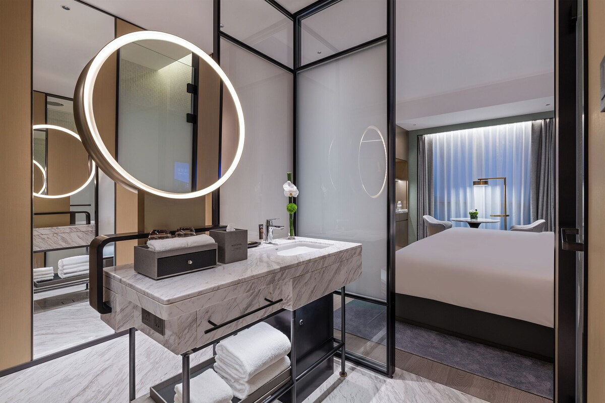 Casa de banho de hotel moderna com toucador em mármore, grandes espelhos e uma cama com vista para a cidade.