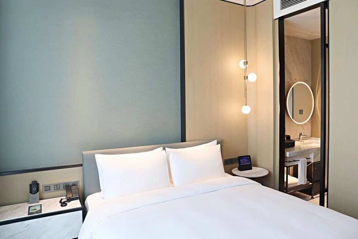 Quarto de hotel com cama, mesa de cabeceira e um banheiro visível através de uma porta aberta.
