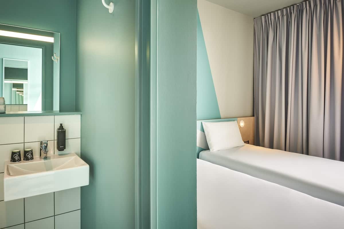 A modern hotel room with a white sink, a mirror, a bed with a white pillow, and a grey curtain.