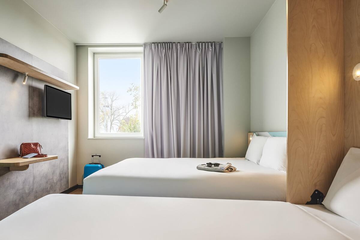 A hotel room with two beds, a TV mounted on the wall, a window with curtains, and a wooden panel on the right side.