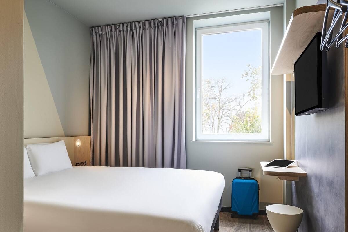 A hotel room with a large bed, a window with curtains, a desk with a computer, a toilet, and a suitcase.