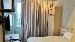 A modern hotel room with a bed, a bedside table, a mirror, a wardrobe, and a window with curtains. - Ibis Budget Nancy Laxou (Laxou)