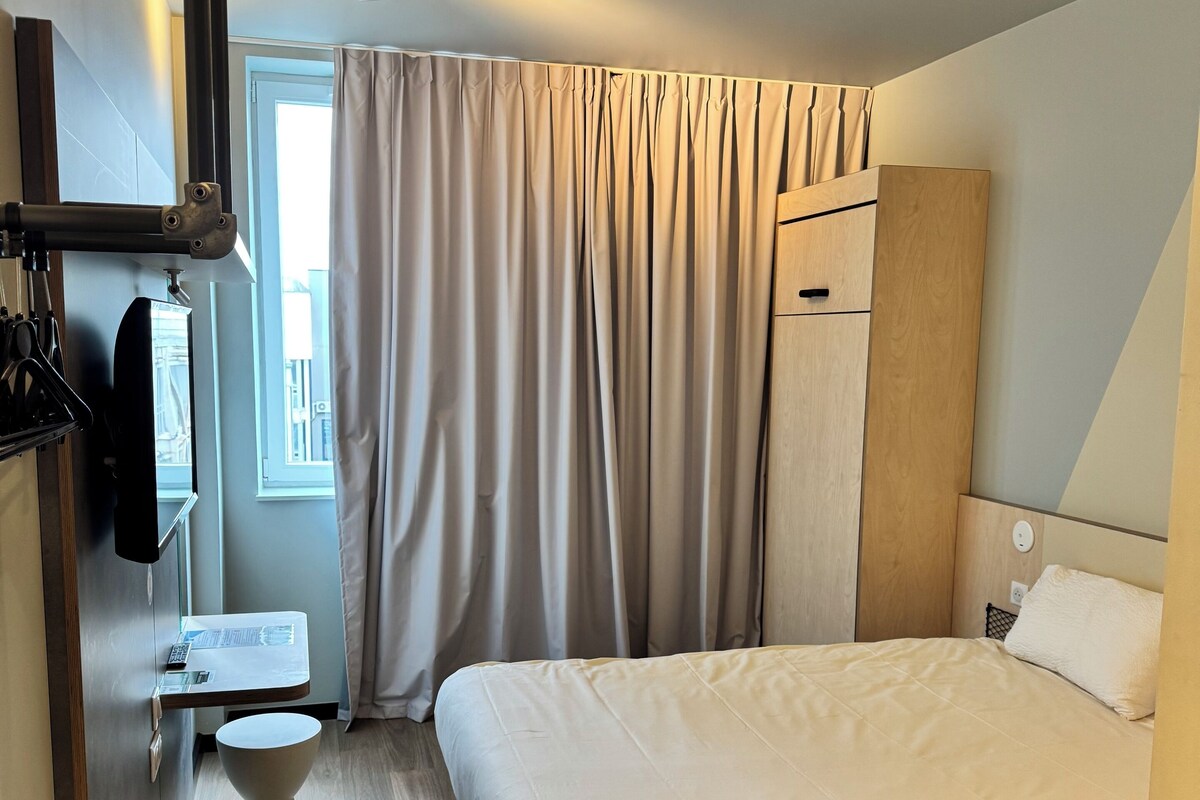 Una habitación de hotel moderna con una cama, una mesita de noche, un espejo, un armario y una ventana con cortinas.