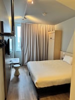 Una habitación de hotel moderna con una cama, una mesita de noche, un espejo, un armario y una ventana con cortinas.