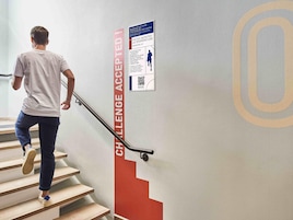 Una persona bajando una escalera con barandilla, un cartel en la pared y un código QR.