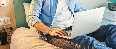 Una persona sentada en una cama con un ordenador portátil, un almohadón y una mesita de noche con una lámpara.