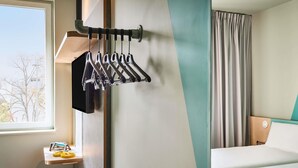 A hotel room with a bed, a wall-mounted coat rack, a small table with toiletries, and a view of trees outside the window. - Ibis Budget Nancy Laxou (Laxou)