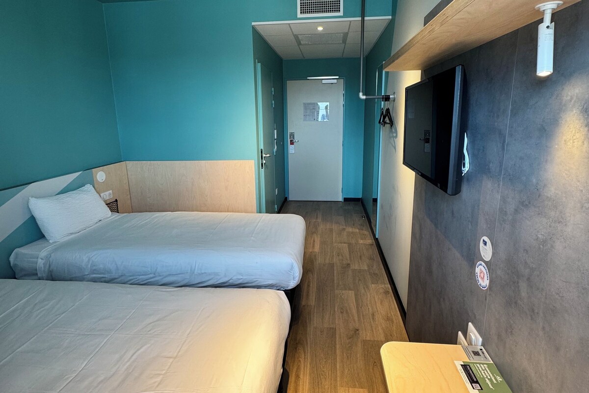 Habitación de hotel con dos camas, suelo de madera, televisor montado en la pared y una mesita con un control remoto.