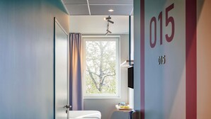 A modern hotel room with a large window, a bed, a bedside table, a chair, and a toilet. - Ibis Budget Nancy Laxou (Laxou)