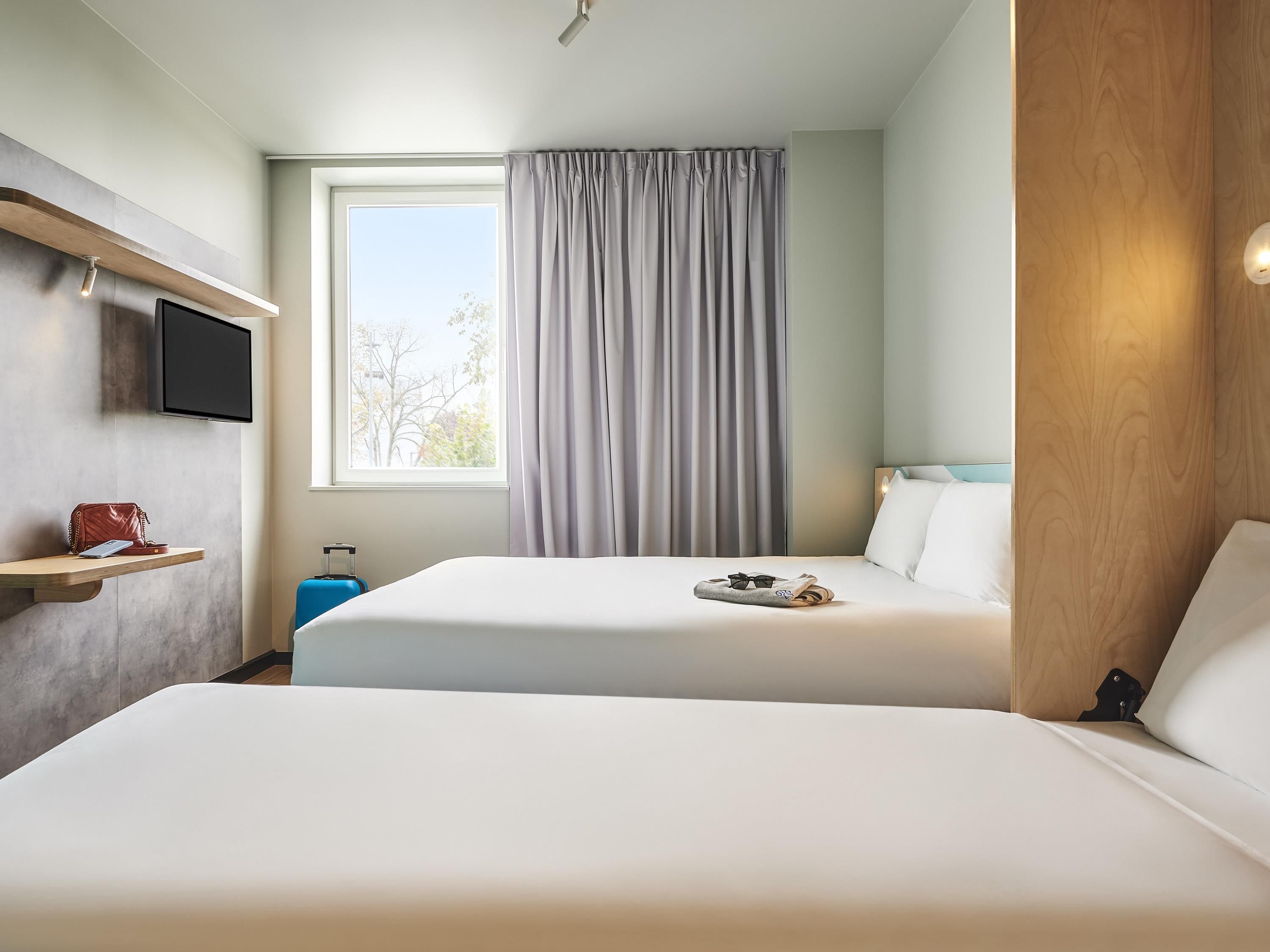 Habitación de hotel con dos camas, un televisor montado en la pared, una ventana con cortinas y un panel de madera a la derecha.