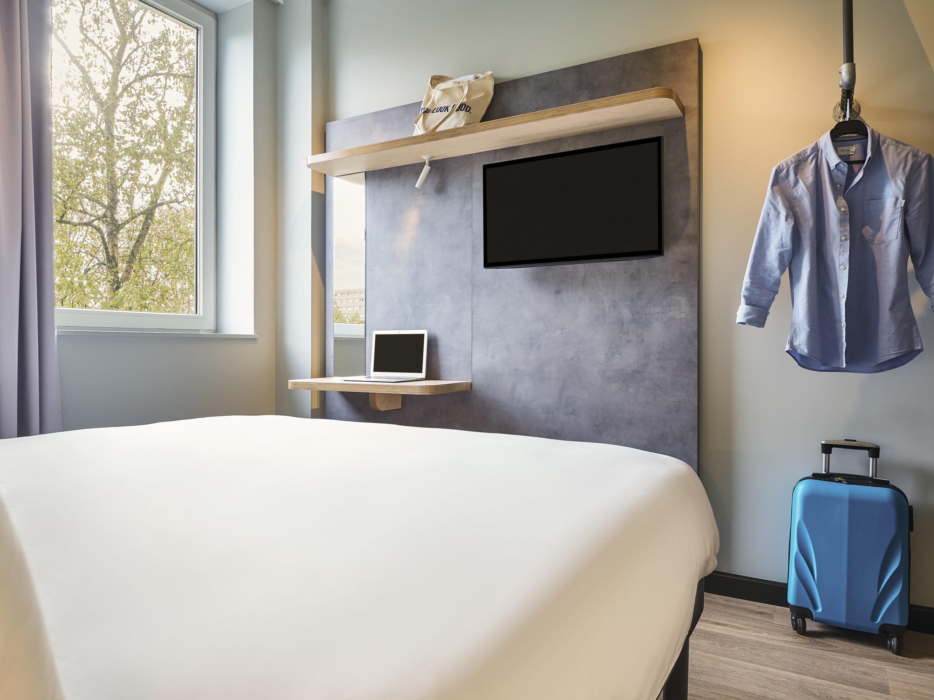 A hotel room with a bed, a desk with a laptop, a TV mounted on the wall, a suitcase, and a window with curtains.