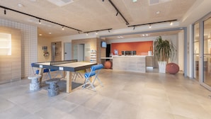 A modern indoor space with a ping pong table, blue chairs, a reception area with a TV, and a large potted plant.