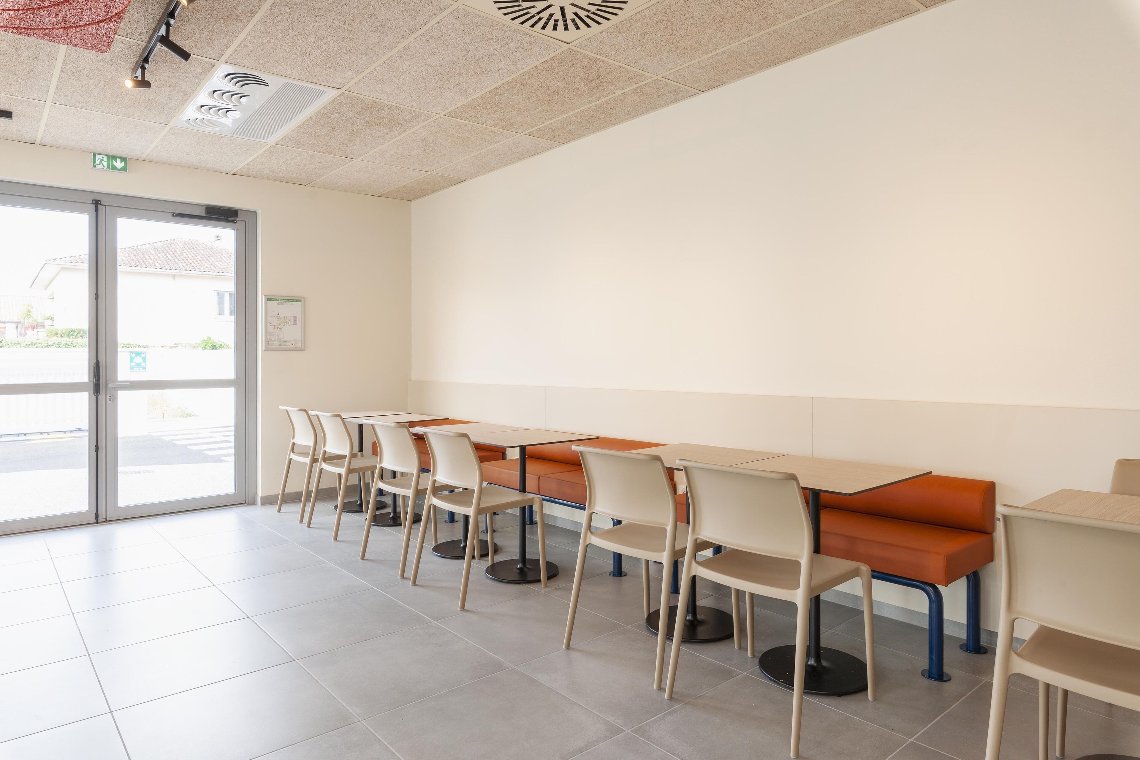 A clean, modern dining area with multiple tables and chairs, a glass door entrance, and a ceiling with ventilation.