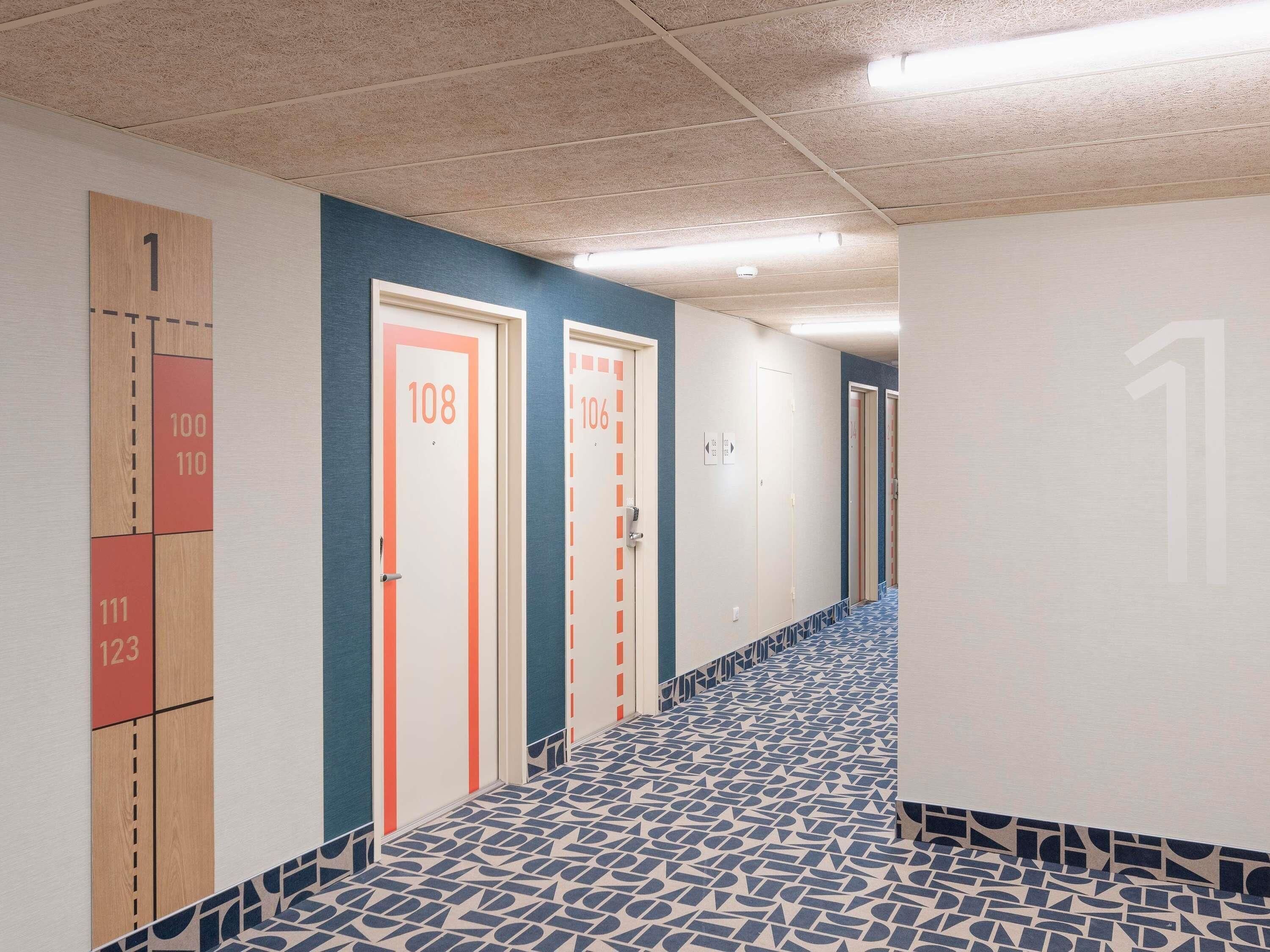 Un pasillo con puertas numeradas, baldosas de suelo con diseño y techo con luces fluorescentes.