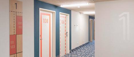 A hallway with numbered doors, patterned floor tiles, and a ceiling with fluorescent lights.