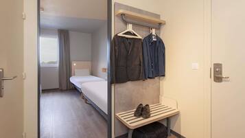 A hotel room with a bed, a bench with shoes and a bag, a coat rack with clothes, and a door.