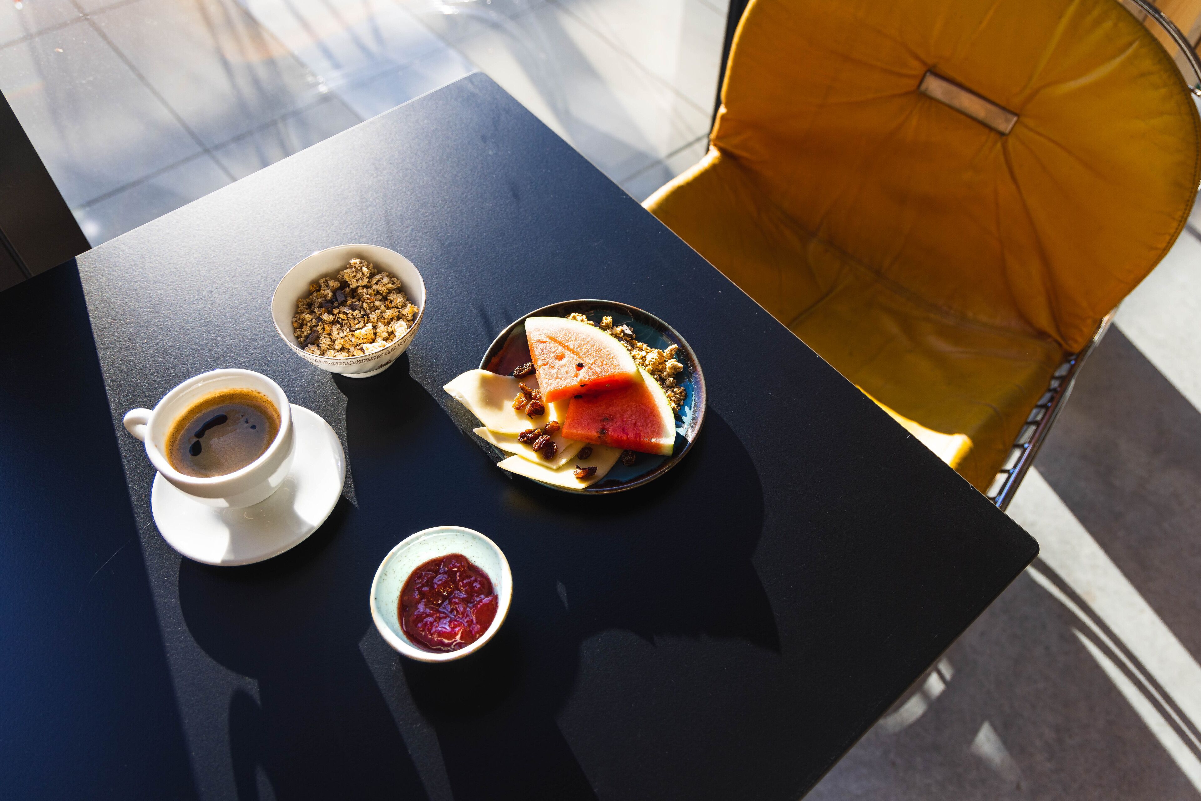 Une table avec une tasse de café, des bols de granola et de confiture, et une assiette avec une tranche de pastèque.