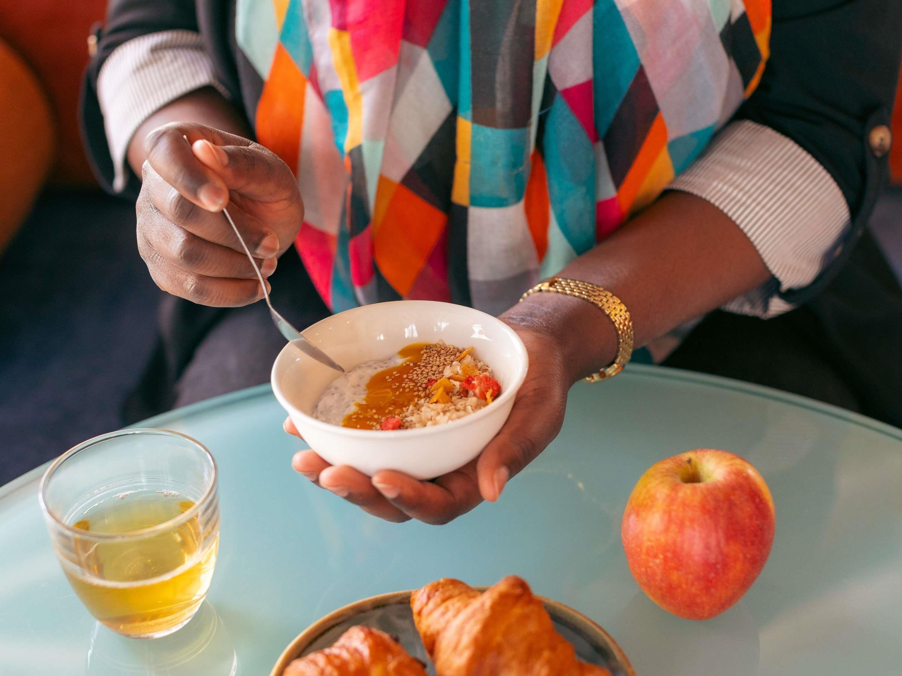 Una persona sosteniendo un tazón de cereal con una cuchara, una manzana y un vaso de jugo sobre una mesa.