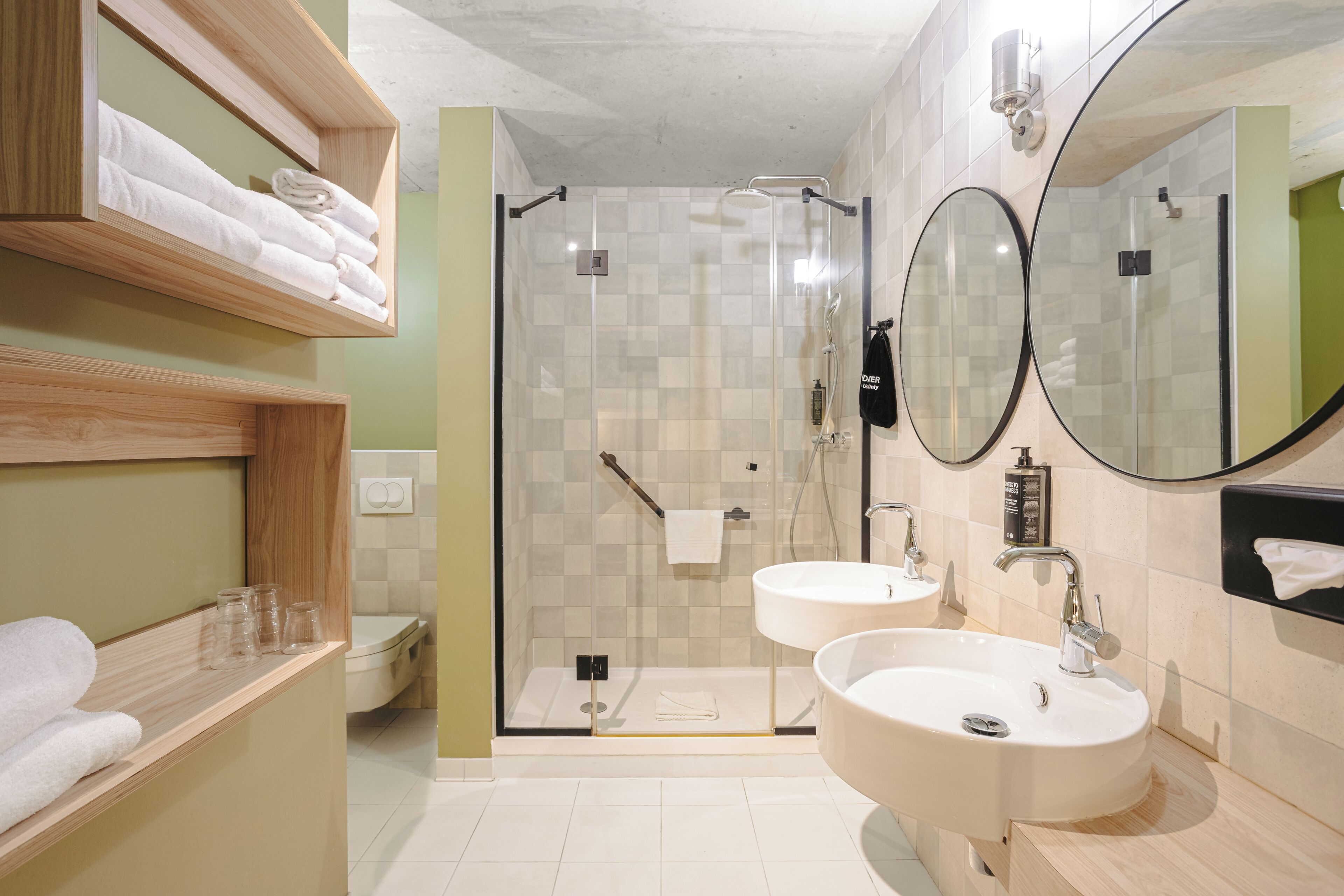 A modern bathroom with two sinks, a glass shower enclosure, and a wooden shelf with towels.