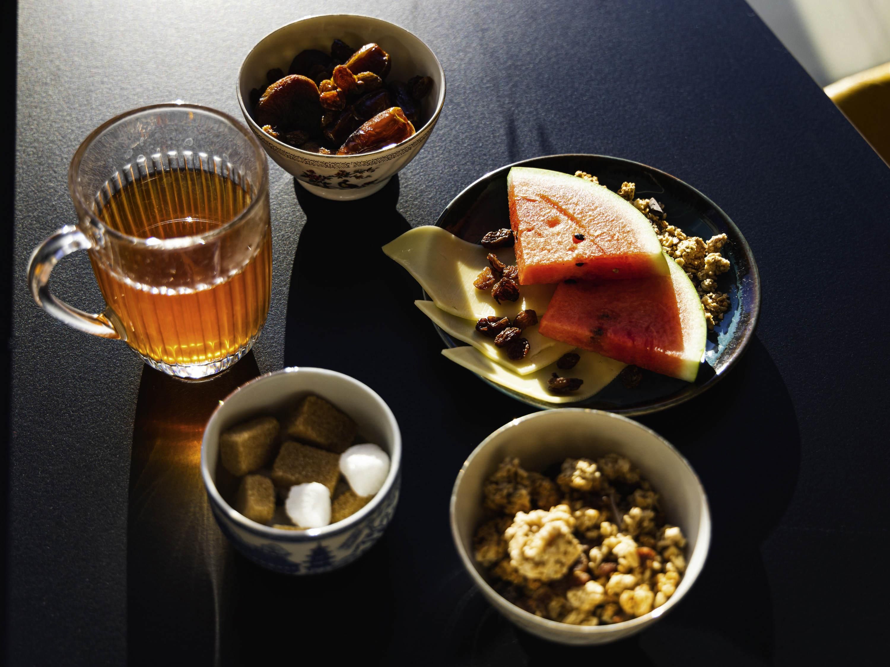 A table with a glass of amber liquid, bowls of food, and a plate with a slice of watermelon and nuts.