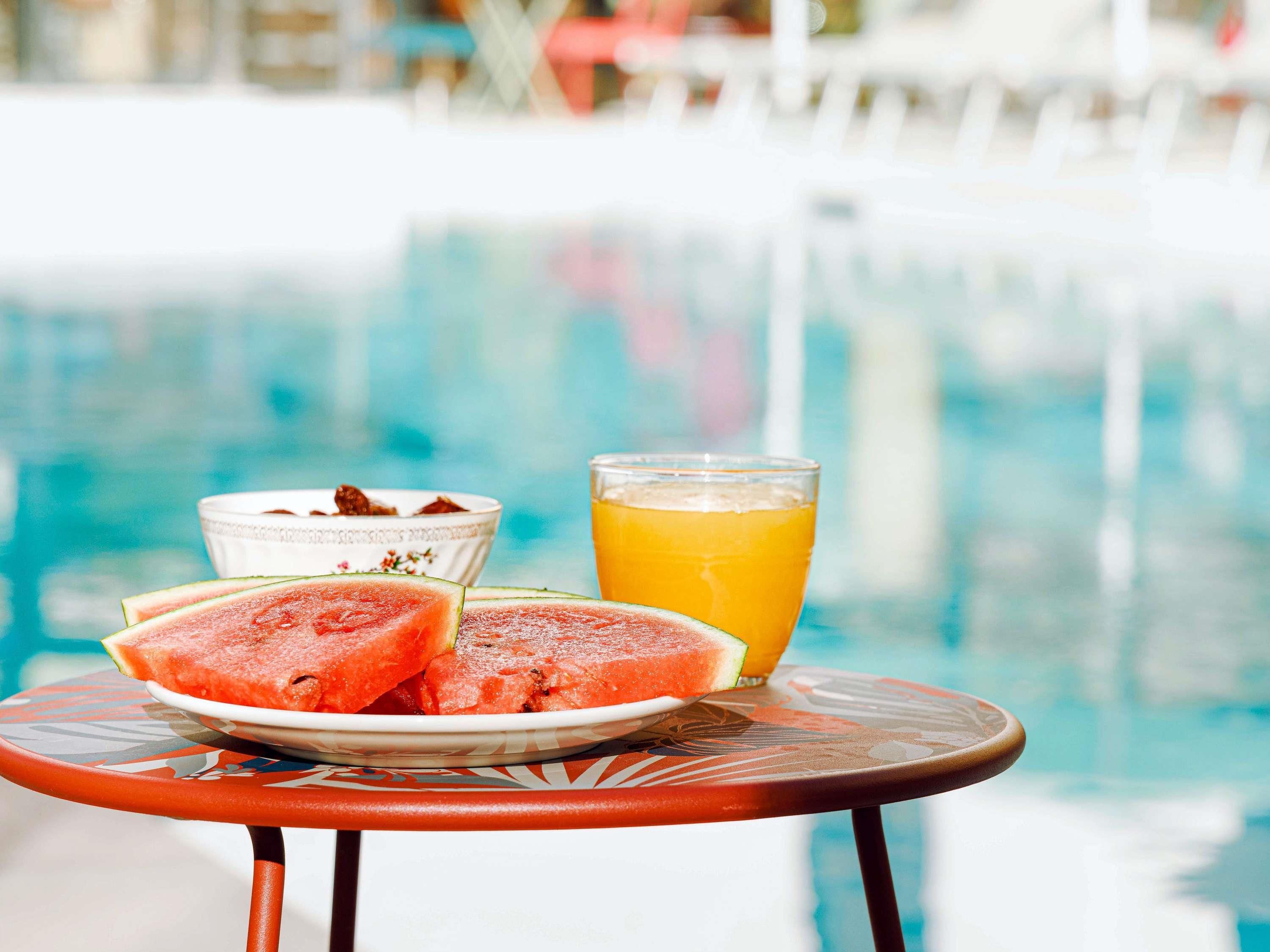 A poolside table with a plate of watermelon slices, a glass of orange juice, and a small bowl of yogurt.