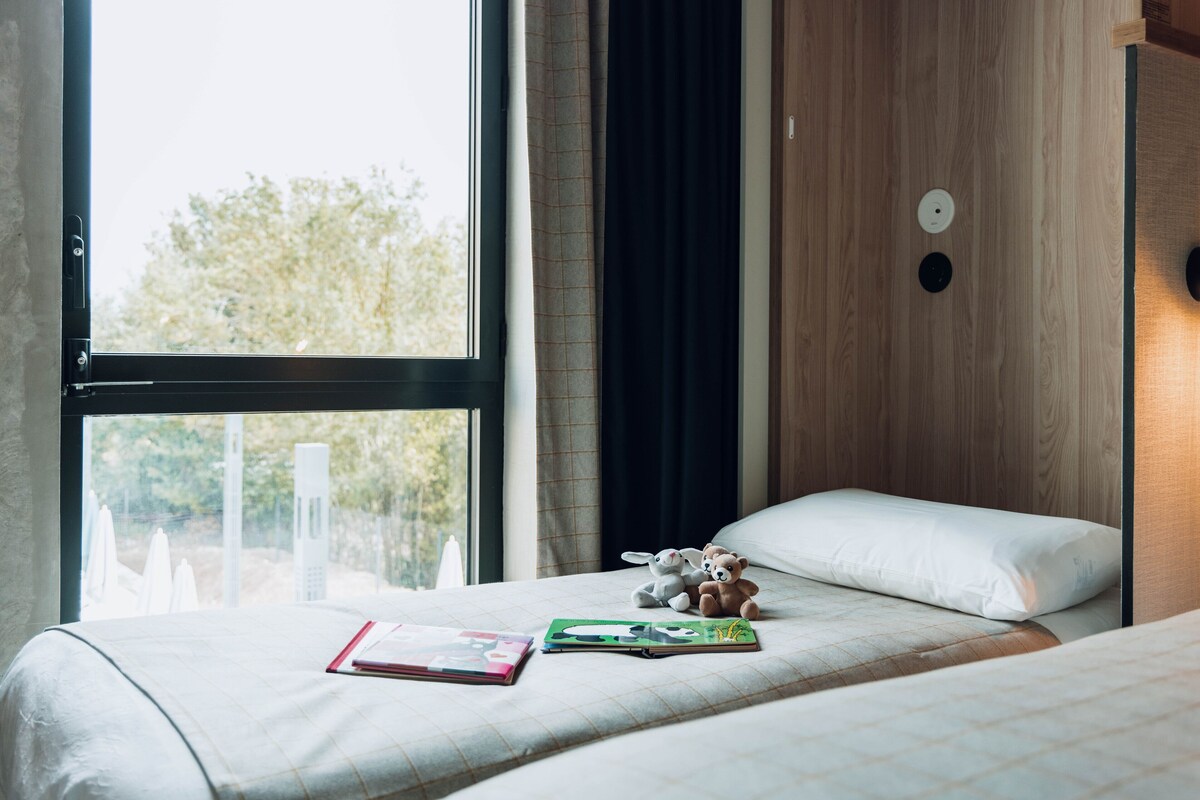 Ein Hotelzimmer mit großem Fenster, einem Bett mit weißem Kopfkissen und einem Nachttisch mit Buch und Teddybär.